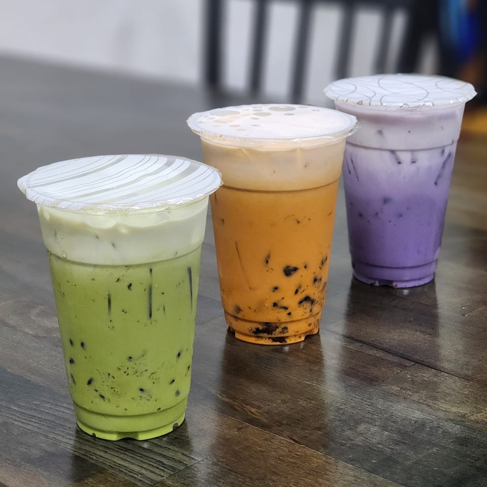 Three colorful bubble tea drinks from Pho Vy: green, orange, and purple, on a wooden table.